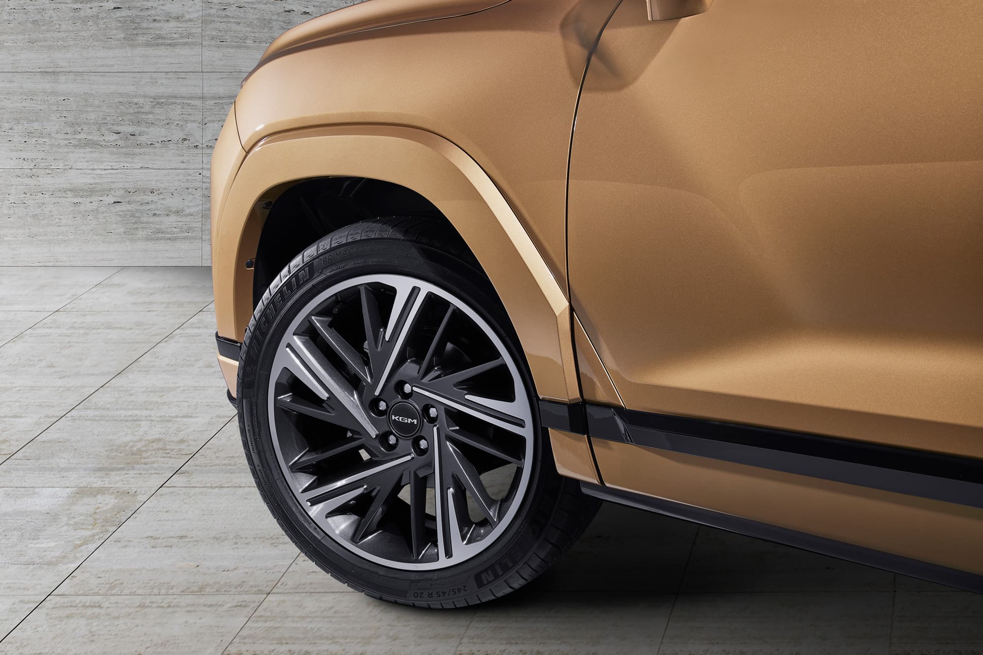Close-up of a bronze SUV's front wheel and tire on a tiled floor, highlighting the vehicle's sleek design and modern alloy wheel.