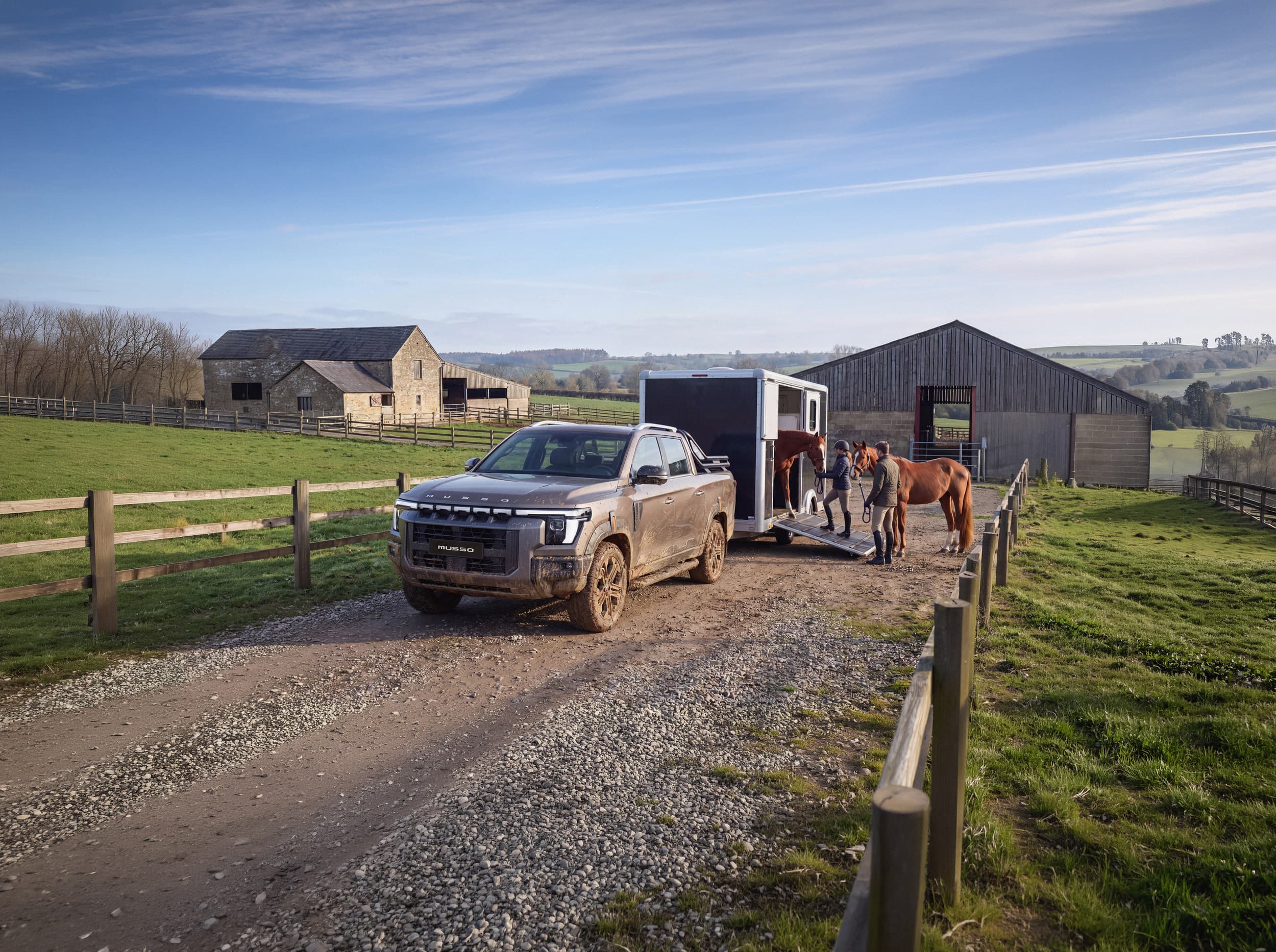 A muddy KGM Musso with a trailer is parked on a farm road as two people load horses. Barns and fields are in the background under a blue sky.