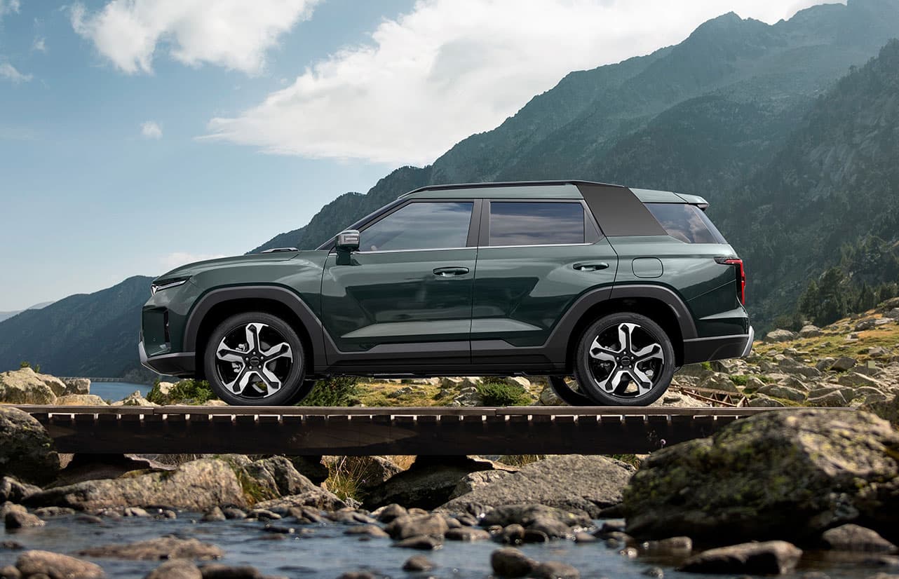 A green KGM Torres Hybrid parked on a wooden bridge over a stream, with mountains and a partly cloudy sky in the background.