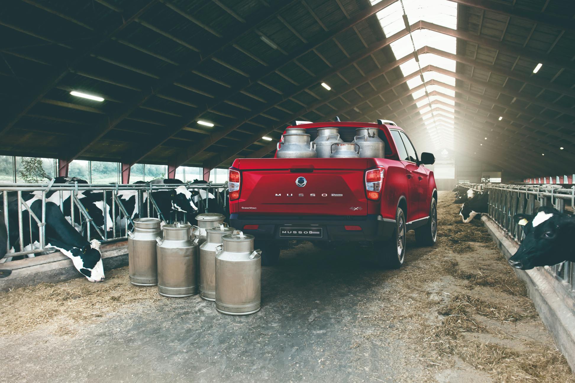 Een rode Grand Musso pick-up, geparkeerd in een boerderij waarbij de laadbak volgeladen is met melkkannen.