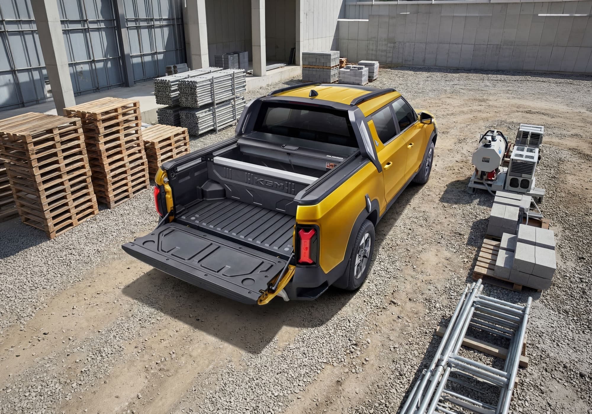 Yellow KGM Musso EV with its tailgate down, parked in an industrial area surrounded by metal racks, pallets, and stacked concrete blocks.