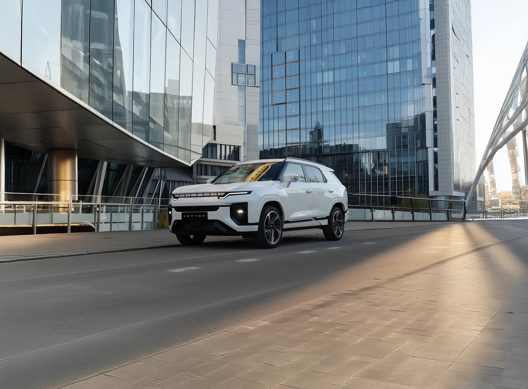 White KGM Actyon Hybrid parked on an empty city street in front of reflective glass skyscrapers and a modern pedestrian bridge.