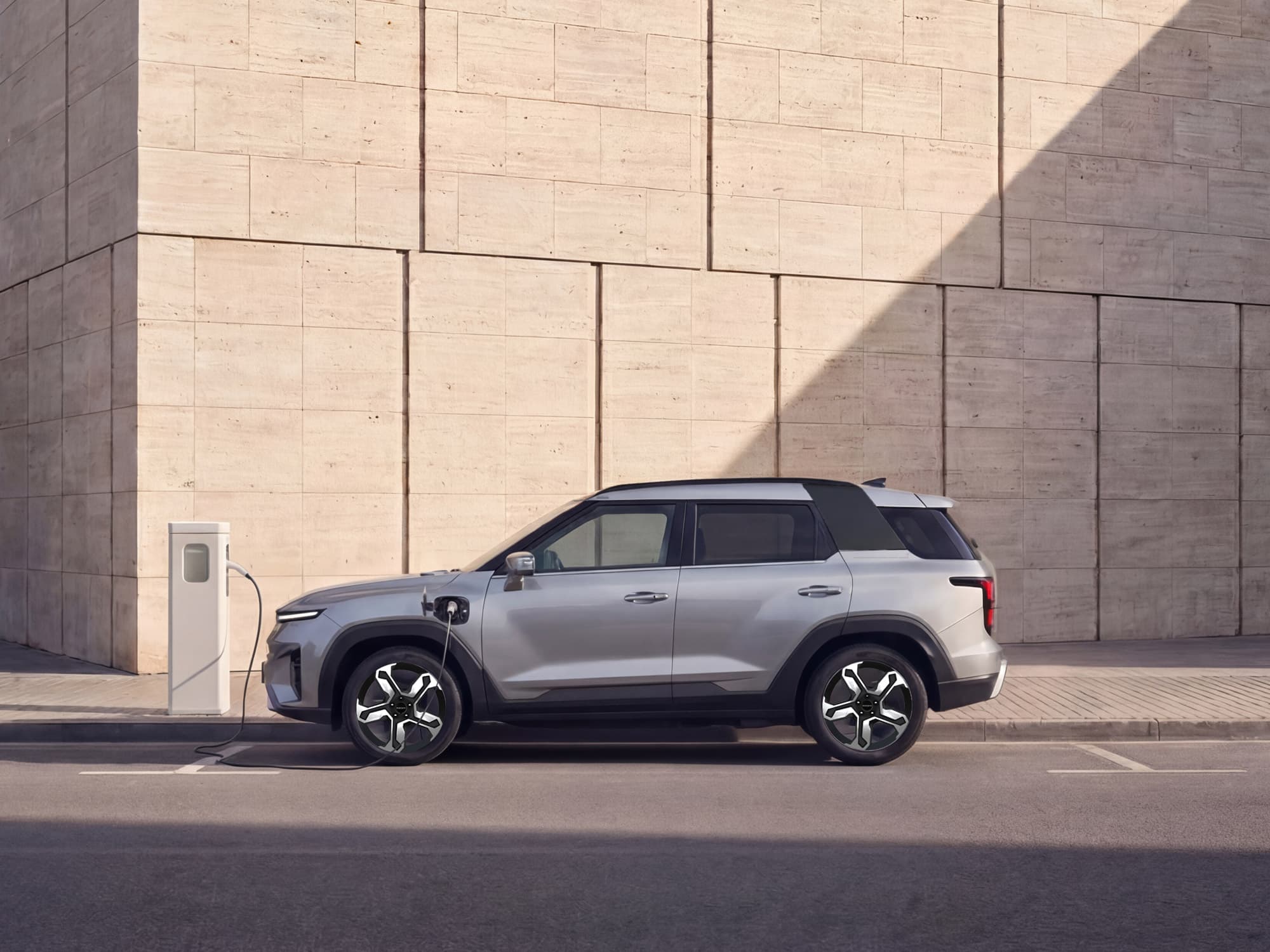 A silver electric Torres EVX is parked and charging at a street charging station against a backdrop of large stone tiles.