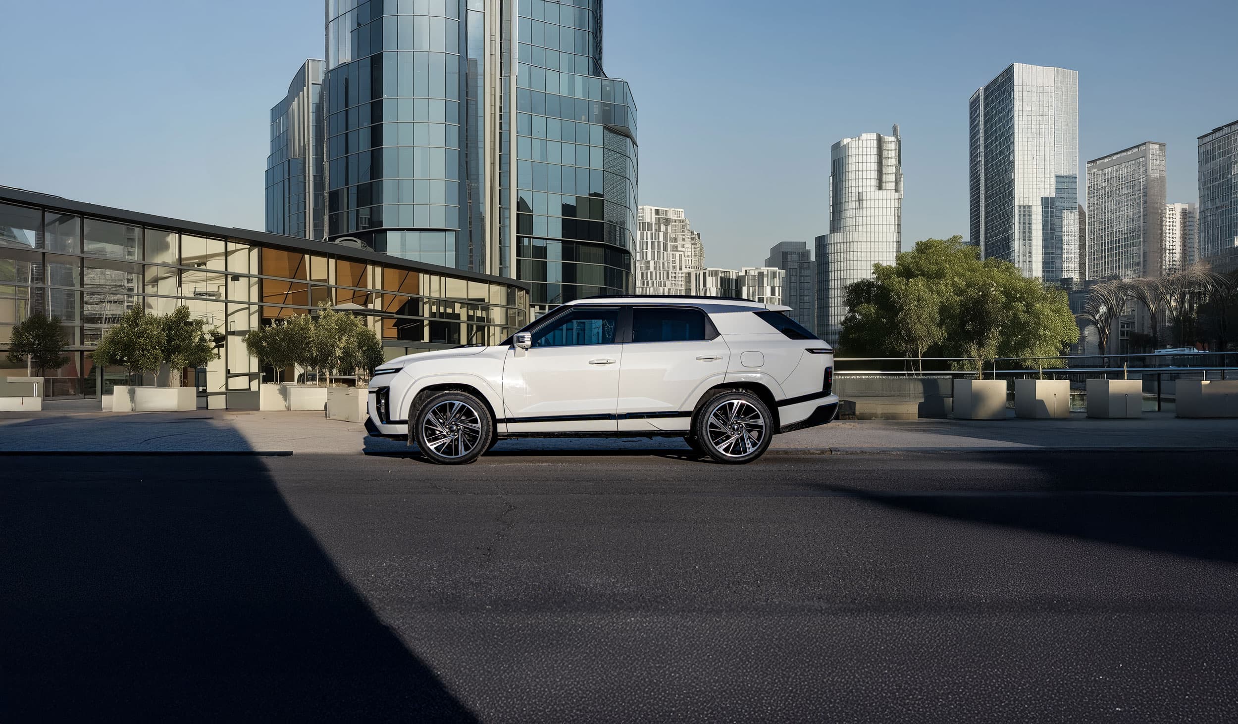 A white KGM Actyon parked on a city street with modern glass buildings and trees in the background under a clear blue sky.