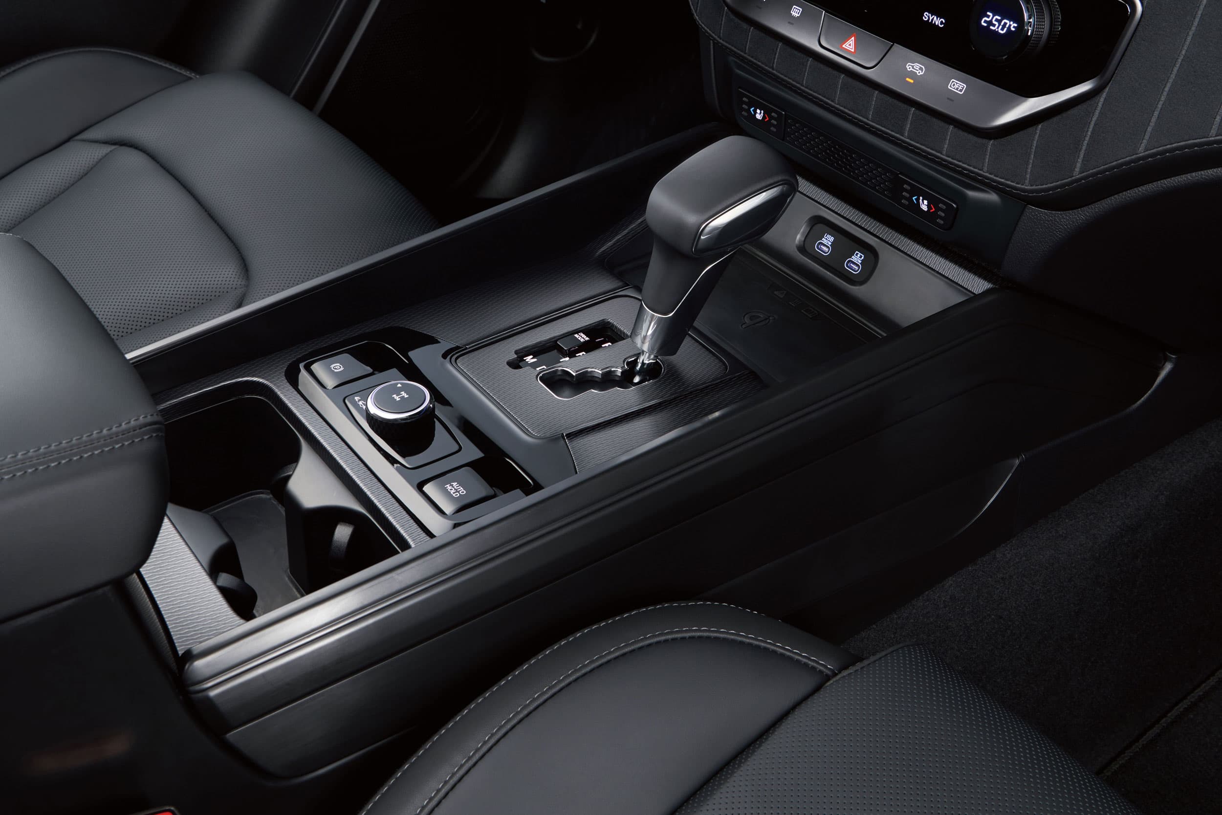 Close-up of a KGM Musso's center console showing a gear shift lever, rotary control knob, cup holders, and climate control buttons on black leather.