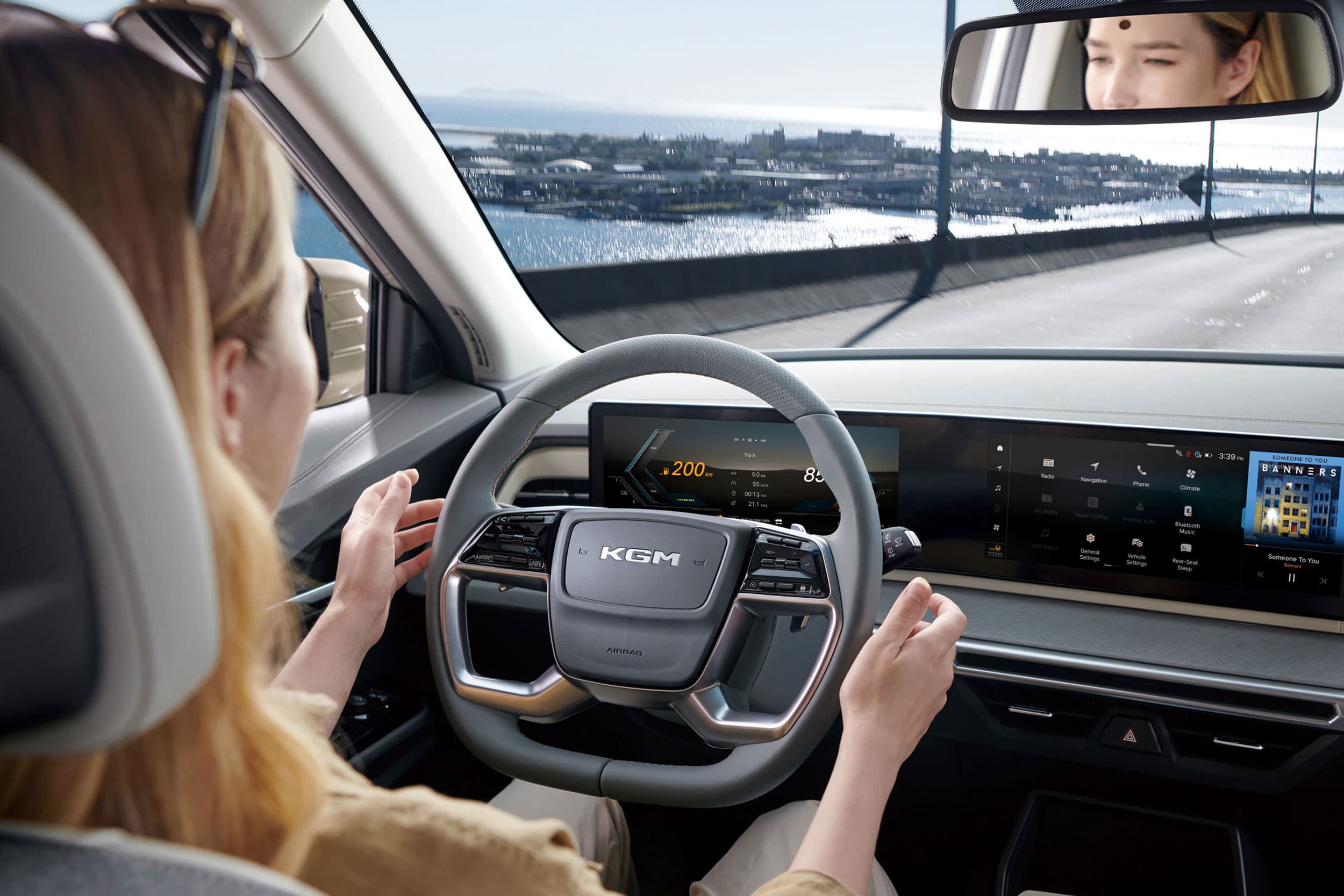 A person driving a KGM Torres Hybrid with a digital dashboard display. The road ahead is clear, and a cityscape and water are visible through the windshield.