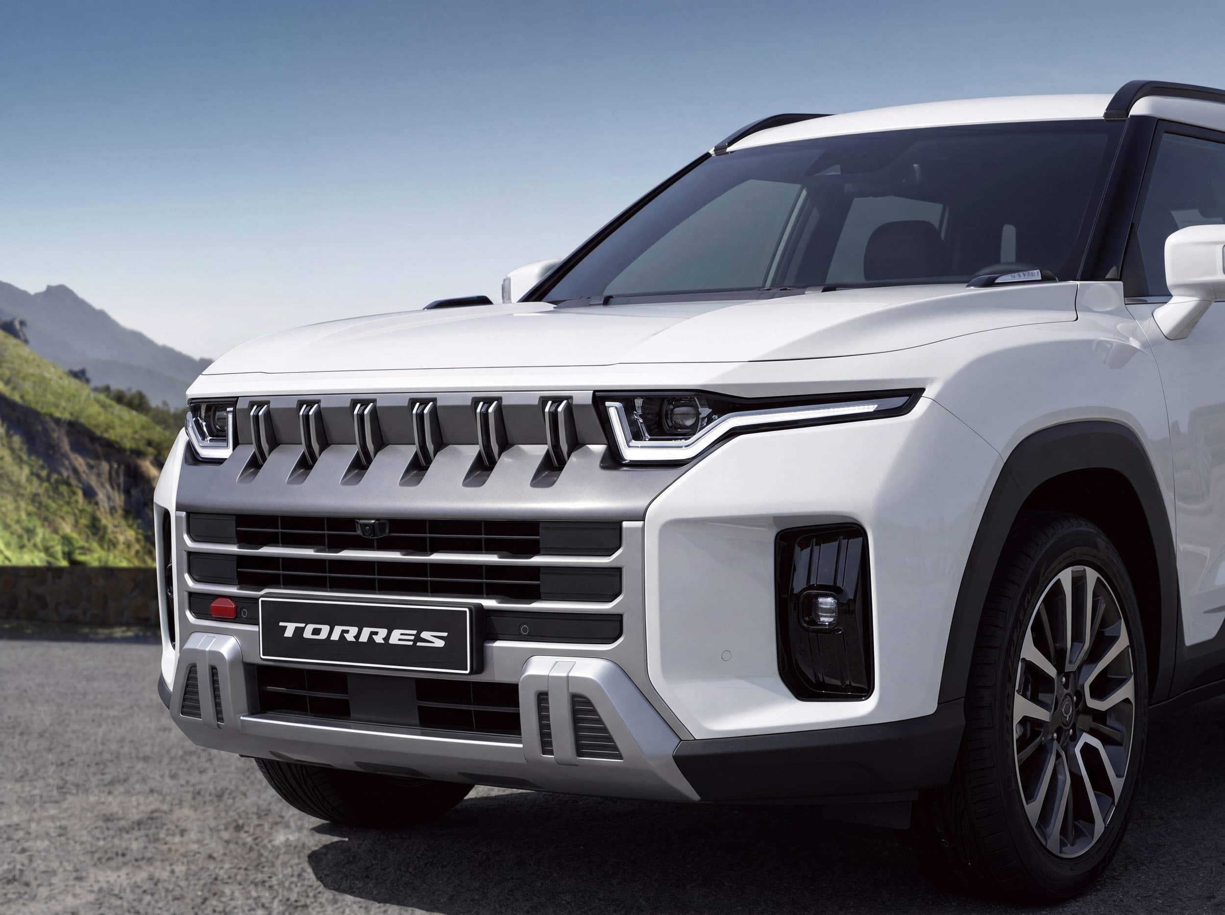 Front view of a white KGM Torres Hybrid with angular headlights and a bold grille design, parked on a road with mountains in the background under a clear sky.