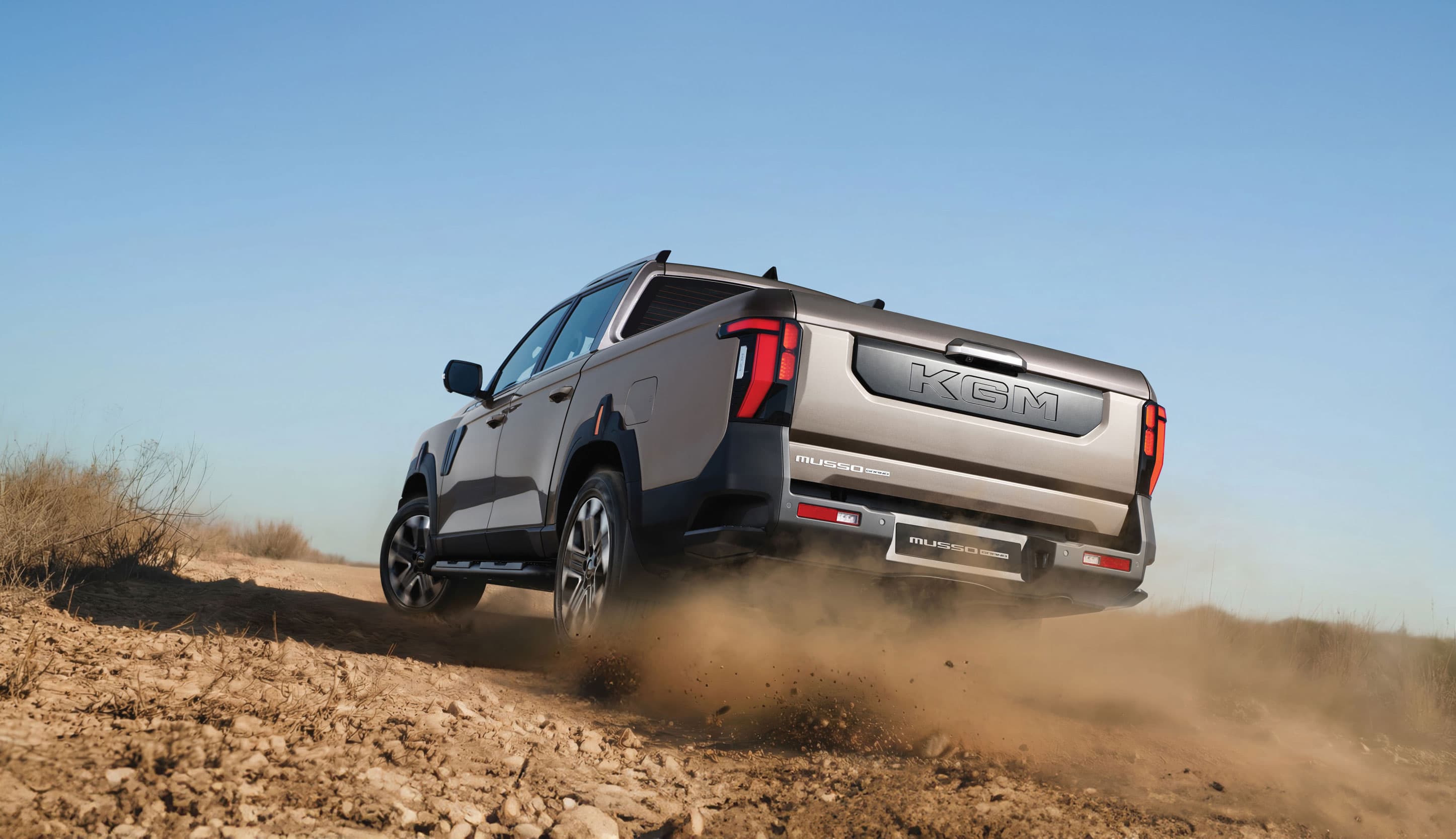 KGM Musso driving on a dusty off-road trail, kicking up dirt, under a clear blue sky.